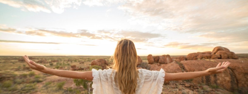 Frau mit ausgebreiteten Armen steht bei Sonnenaufgang in weiter Landschaft – Karfreitag spirituell als Bild von Hingabe und Auferstehung