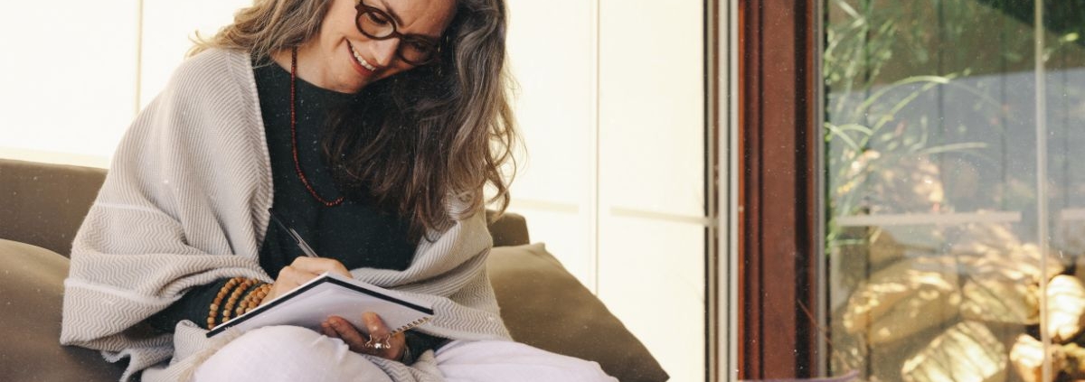 Frau sitzt lächelnd journaling auf dem Sofa und schreibt in ihr Notizbuch. Rauhnächte-Wünsche erfüllen sich nicht sofort, sie entfalten sich über das ganze Jahr
