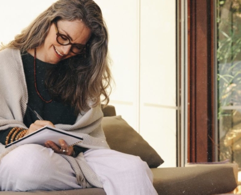 Frau sitzt lächelnd journaling auf dem Sofa und schreibt in ihr Notizbuch. Rauhnächte-Wünsche erfüllen sich nicht sofort, sie entfalten sich über das ganze Jahr
