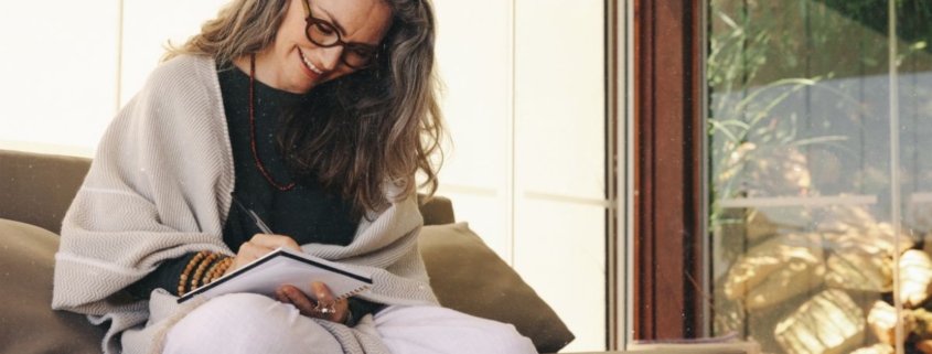Frau sitzt lächelnd journaling auf dem Sofa und schreibt in ihr Notizbuch. Rauhnächte-Wünsche erfüllen sich nicht sofort, sie entfalten sich über das ganze Jahr